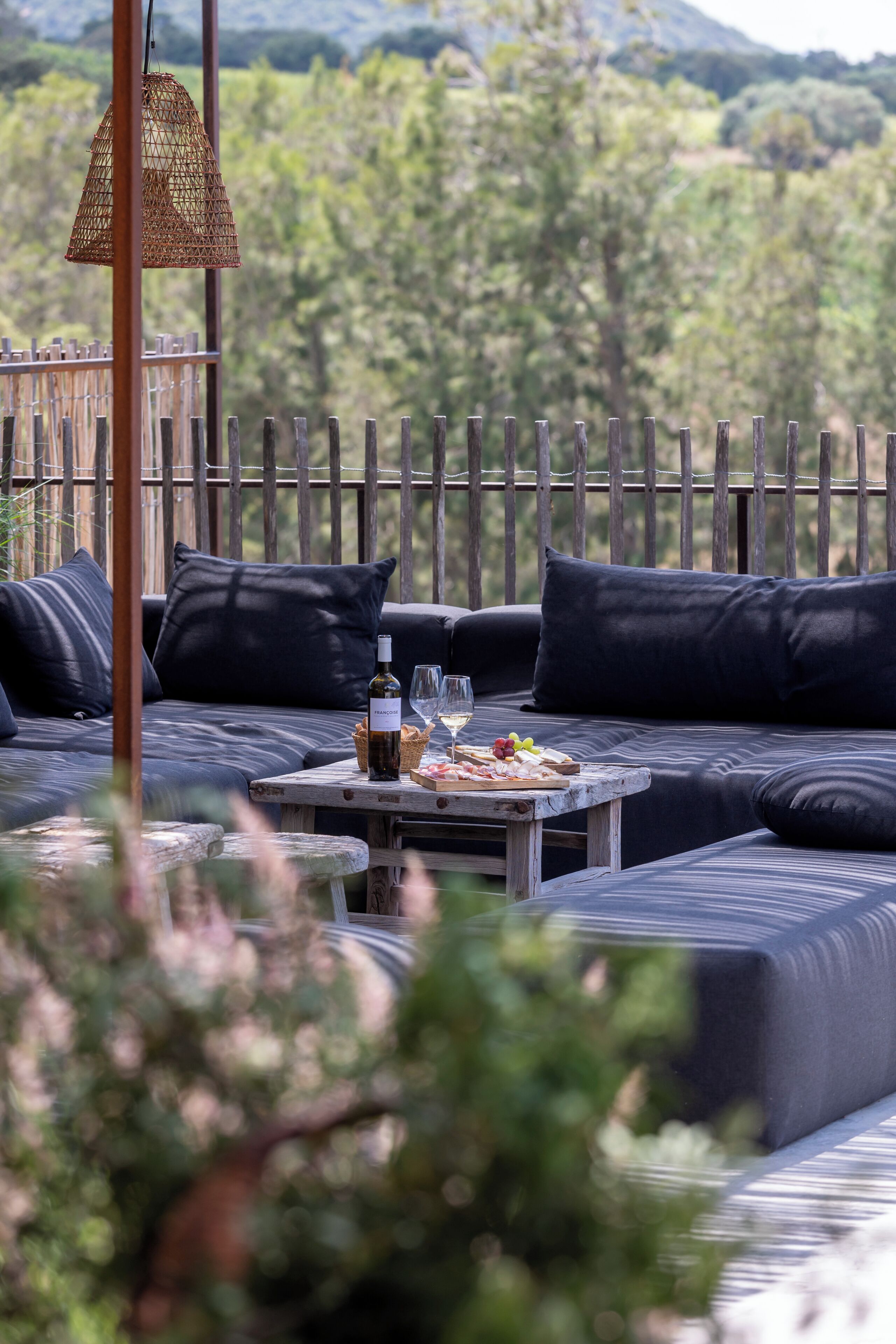 Een terras met een tafel, een fles wijn en glazen, omringd door donkere kussens en een houten hek.