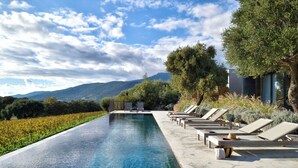 Uma piscina em formato retangular e com borda infinita, cadeiras de descanso e vista para montanhas.