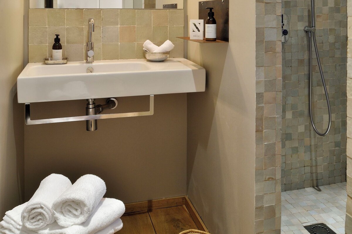 A bathroom with a sink, mirror, towel rack, shower, and a small wooden stool.