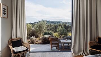 Una habitación con un amplio ventanal corredera que da a un espacio exterior con vistas a árboles y colinas. Hay sillones de mimbre y una mesita con cojín.