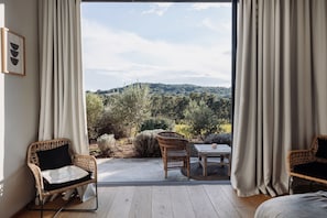 A room with a large sliding glass door leading to an outdoor area with a view of trees and hills. There are wicker chairs and a small table with a cushion.
