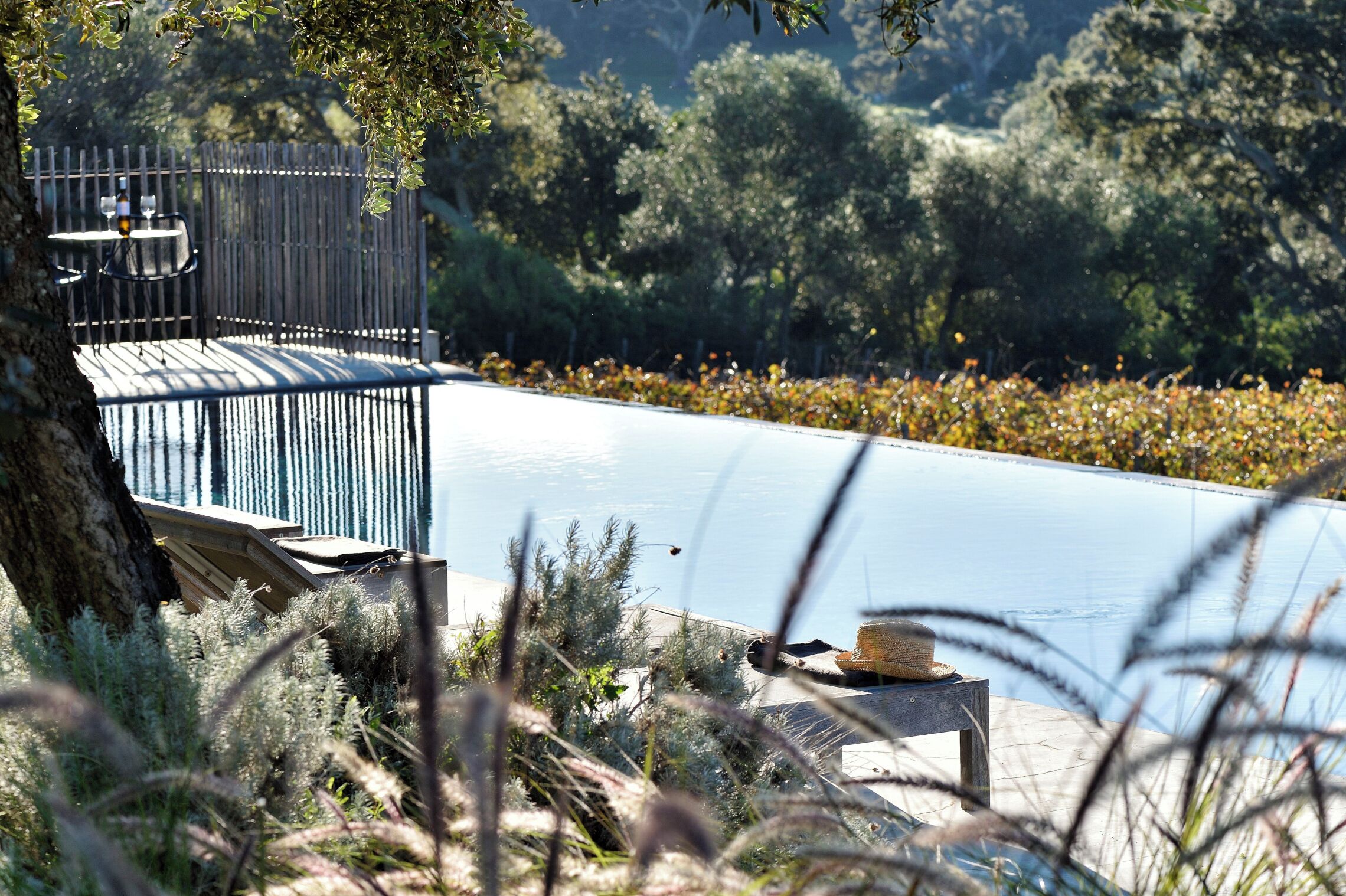 Un área junto a la piscina con una terraza de madera, una mesa con sillas y un sombrero sobre una mesita.