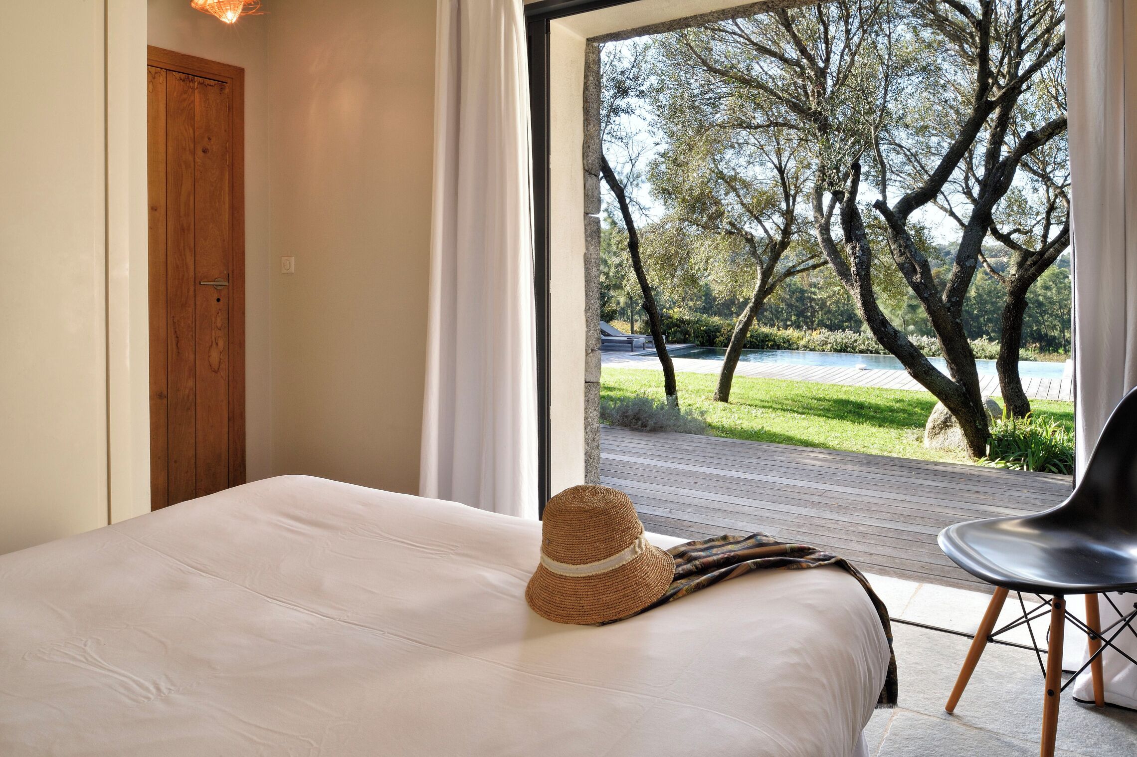 Un dormitorio con cama, una puerta de madera y vistas a un jardín y piscina a través de un ventanal.