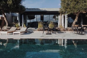 A modern outdoor pool area with lounge chairs, a dining table, and a building with large windows. - Domaine de Peretti della Rocca (Figari)