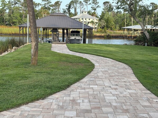 A paved pathway leading to a covered pavilion by a lake, surrounded by well-maintained grass and trees. - Waterfront House with floating Boathouse, can sleep 7. Boat ramp .6 miles (Steinhatchee)