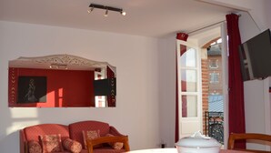 Une pièce chaleureuse avec un canapé rouge, une table à manger recouverte d’une nappe blanche, un saladier blanc et un téléviseur fixé au mur.