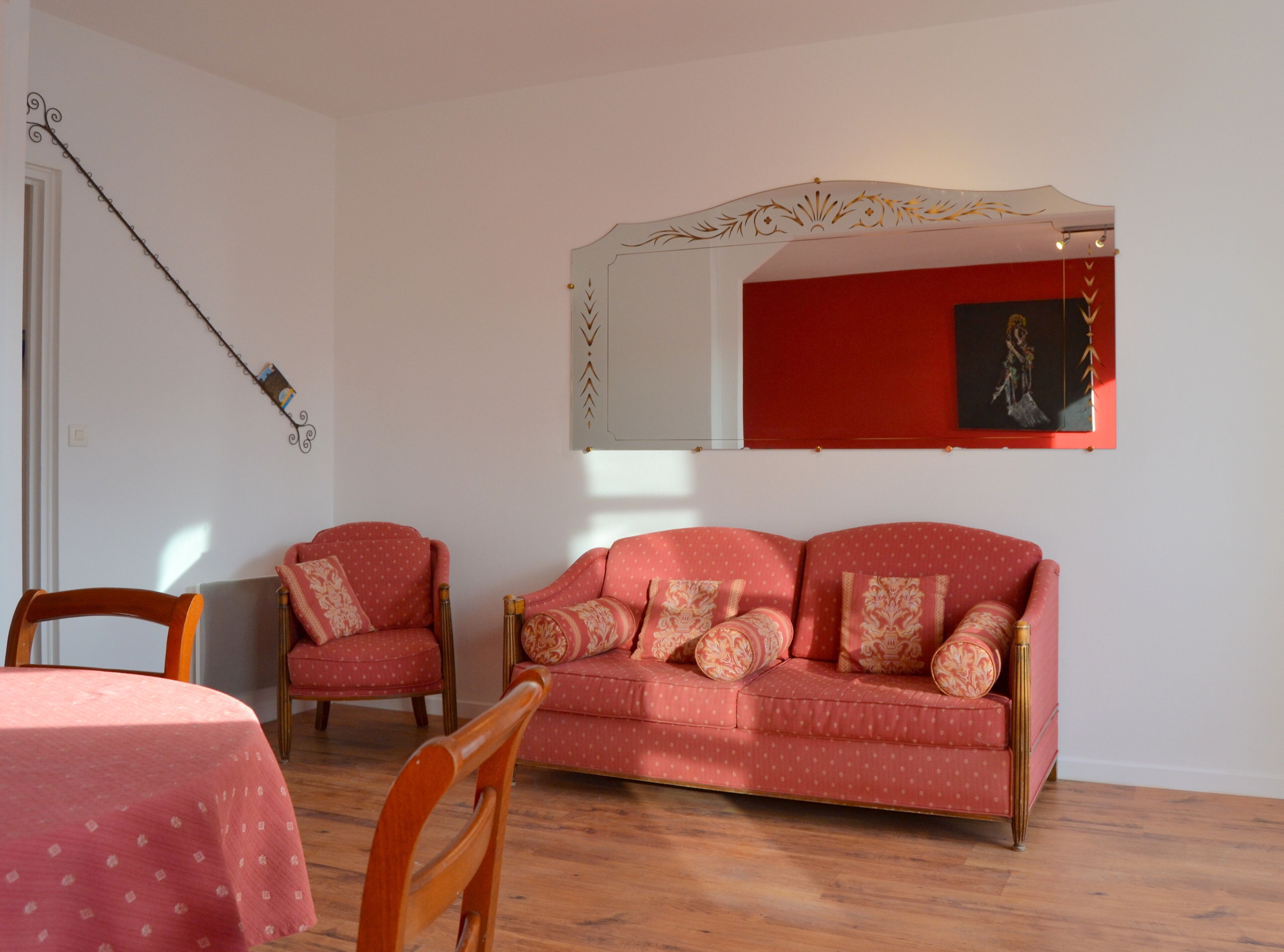 Un salon avec un canapé rouge, deux fauteuils, une table à manger en bois recouverte d’une nappe rose et un miroir fixé au mur.