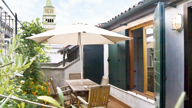 Une terrasse avec une table et des chaises, un grand parasol et une vue sur un bâtiment doté d’un clocher.