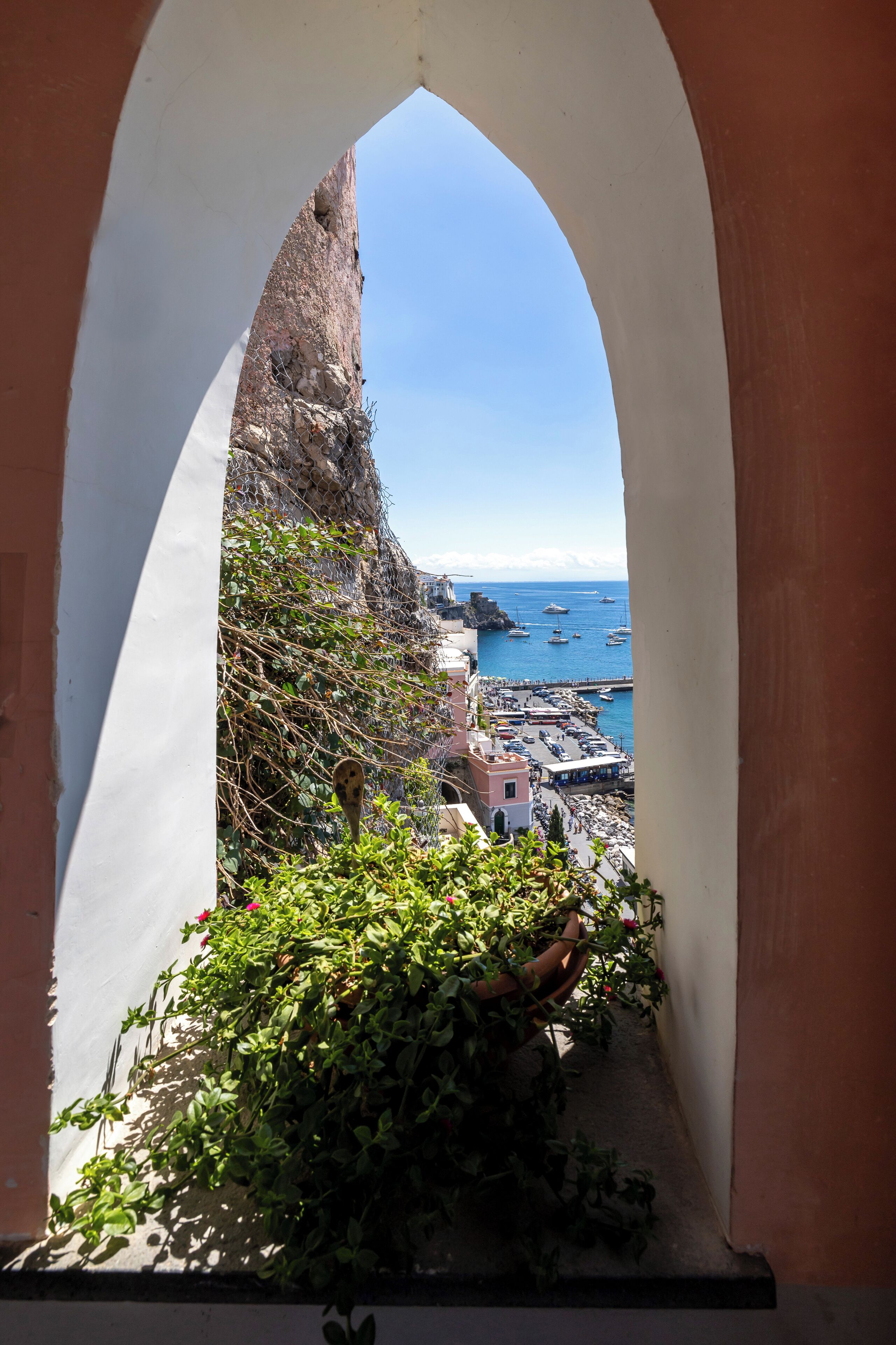 Visão através de um arco que se abre sobre uma cidade costeira, com edifícios, um penhasco rochoso e barcos na água.
