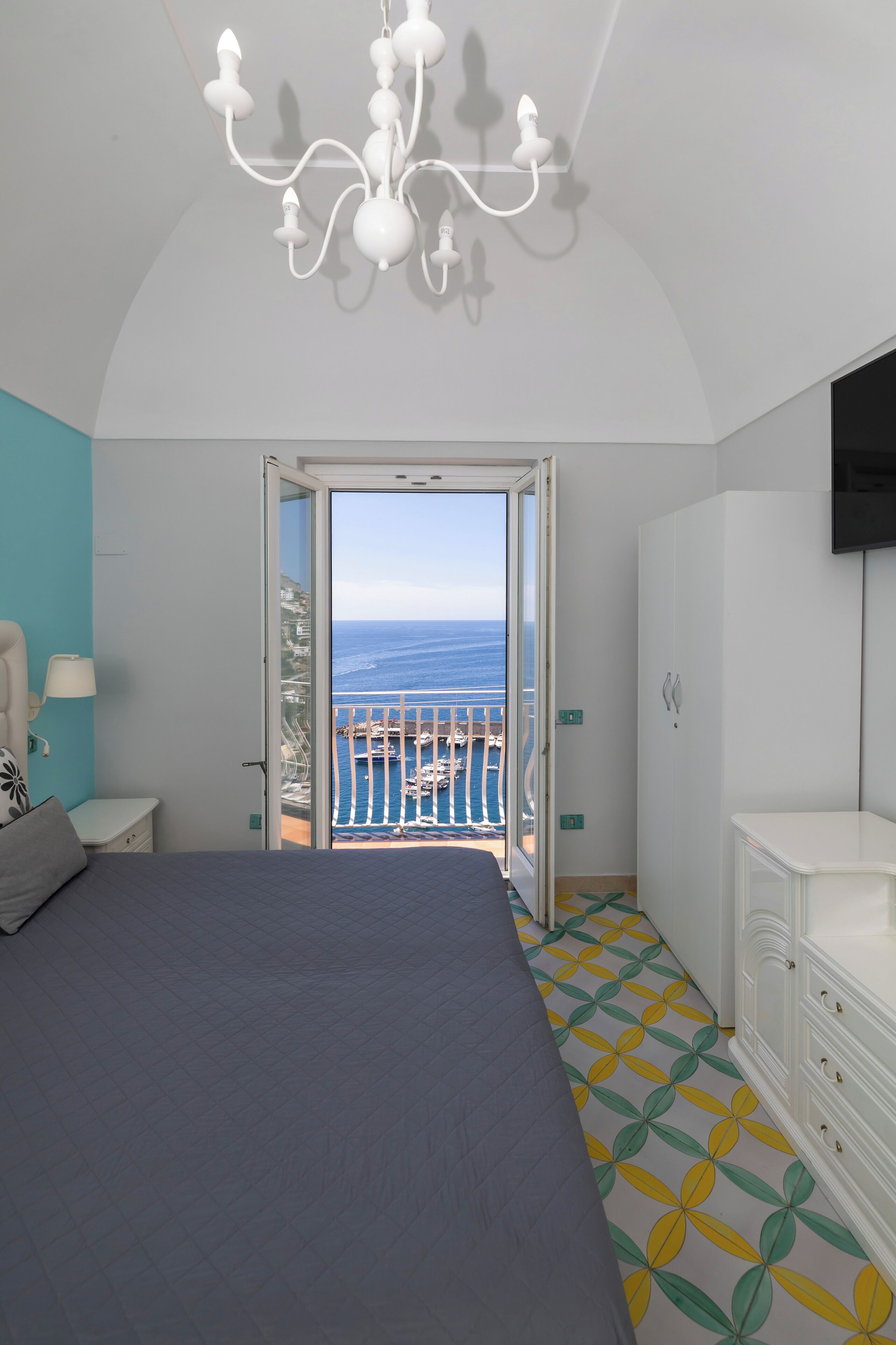 A bedroom with a bed, a chandelier, a wardrobe, and a view of the sea through an open door.