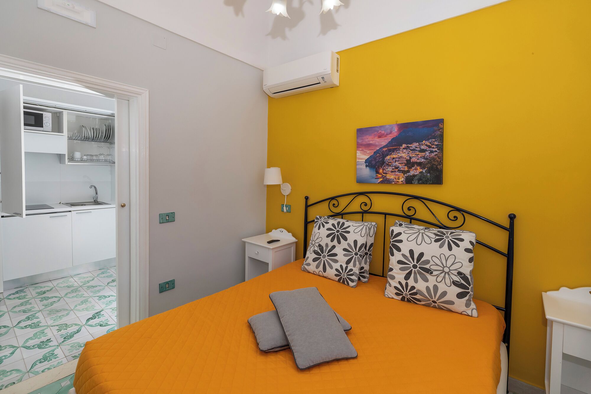 A bedroom with an orange bedspread, a black metal headboard, and two pillows with a floral pattern. There is a painting of a coastal town on the wall.