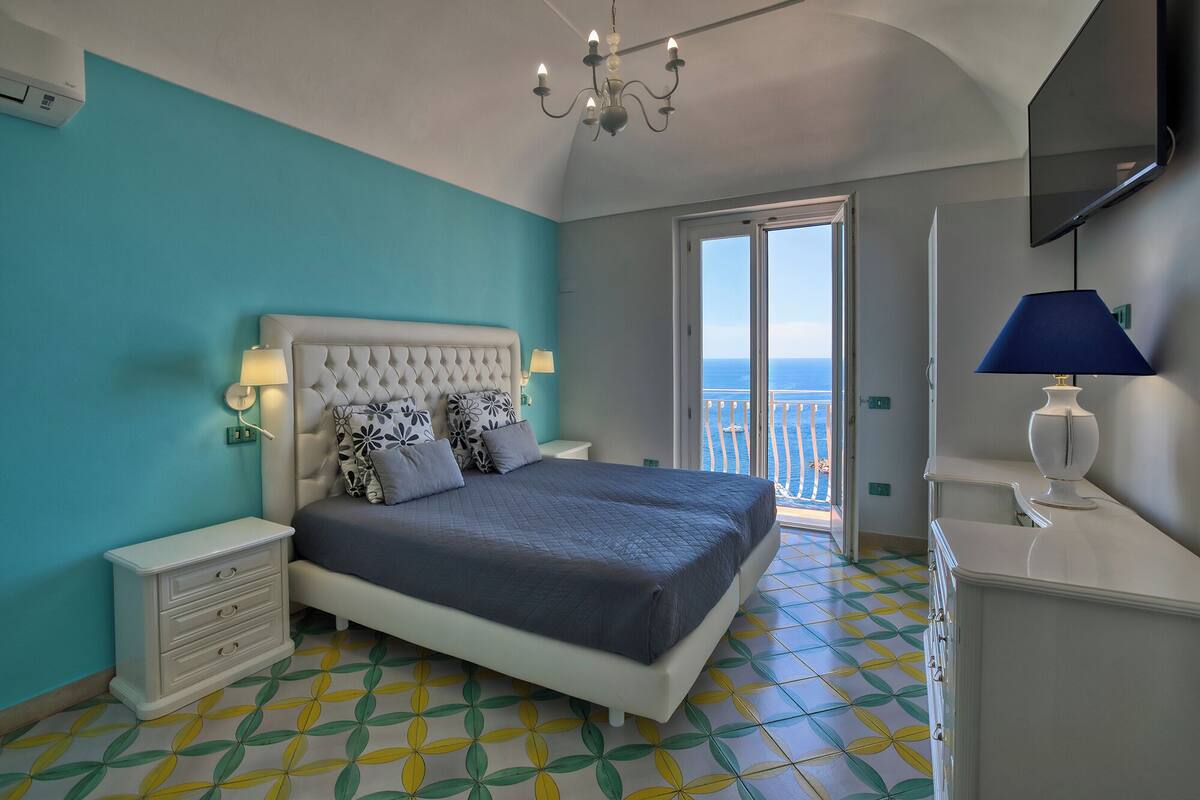 A bedroom with a bed, a dresser, a lamp, a TV, and a view of the sea through a balcony door.