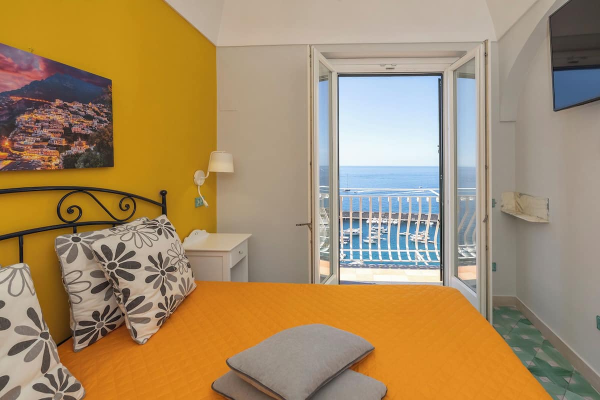 A bedroom with an orange bedspread, a view of a marina through an open door, a painting of a coastal town, and a white nightstand.