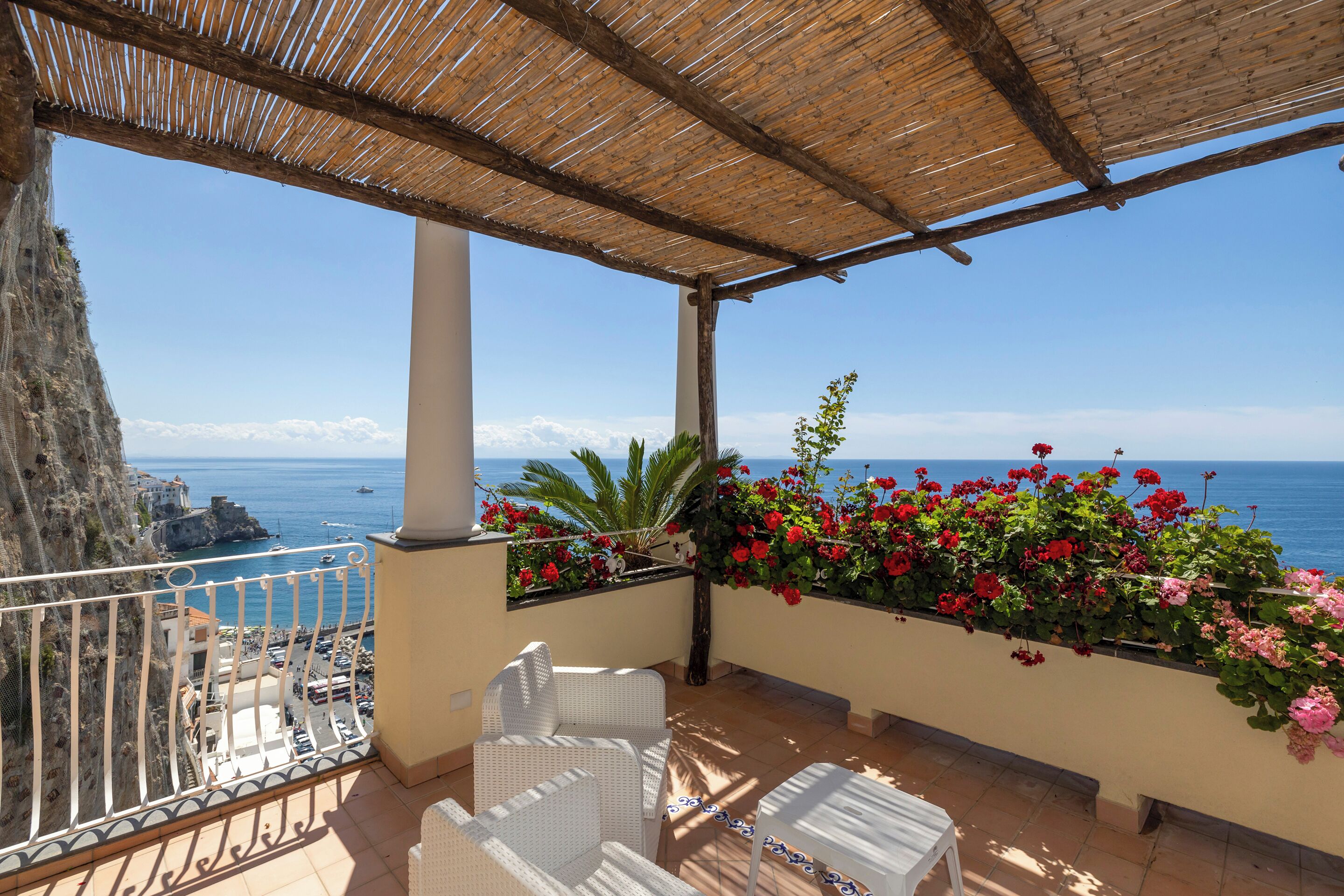 Un balcon agrémenté de meubles en osier, une vue sur la mer et une jardinière remplie de fleurs rouges.