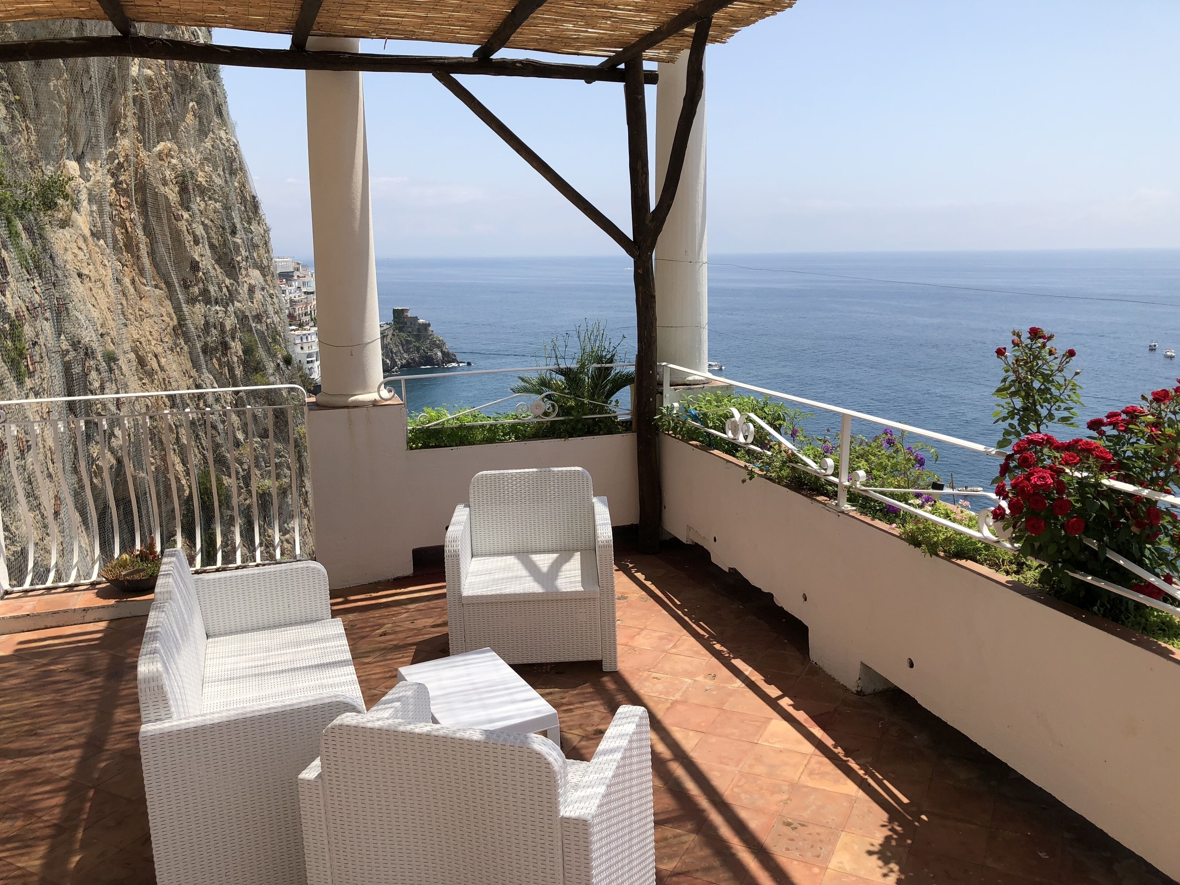 A terrace with white wicker furniture, a view of the sea, and a rocky cliff.