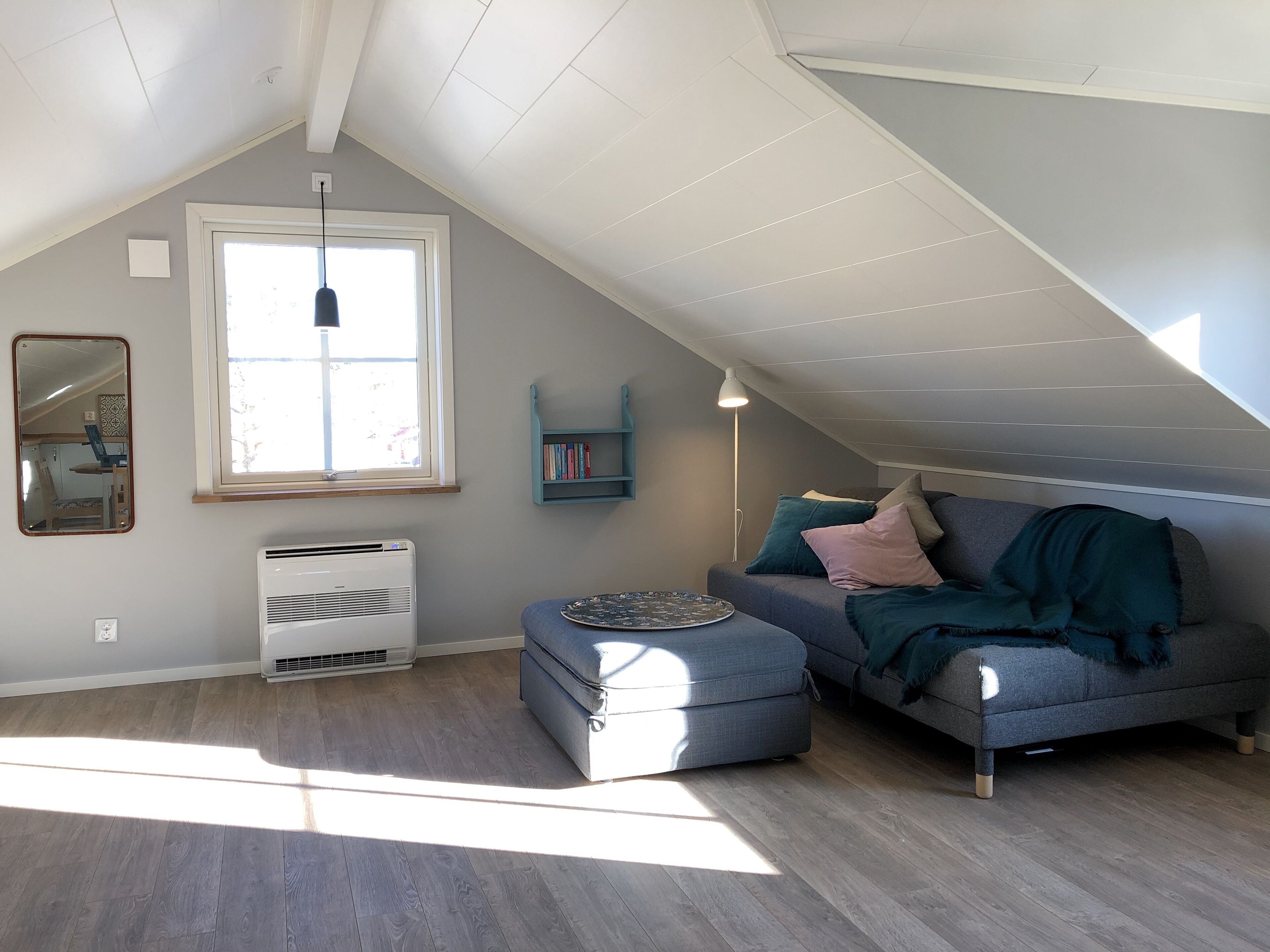 Una habitación con techo inclinado, ventana, sofá, reposapiés, lámpara de pie y estantería.