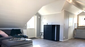 Een moderne loft slaapkamer met een grijze bank, een klein voetenbankje, een zwarte garderobe en een raam dat natuurlijk licht binnenlaat.
