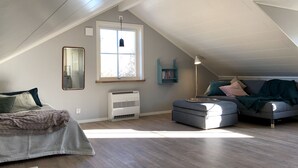 Een knus loft slaapkamer met een schuine kap, een groot raam, een ingebouwde airconditioner, een bedlamp en een gezellige zithoek met kussens en een plaid.