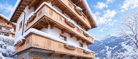 A multi-story wooden chalet with balconies, stone foundation, and a snowy mountain backdrop.
