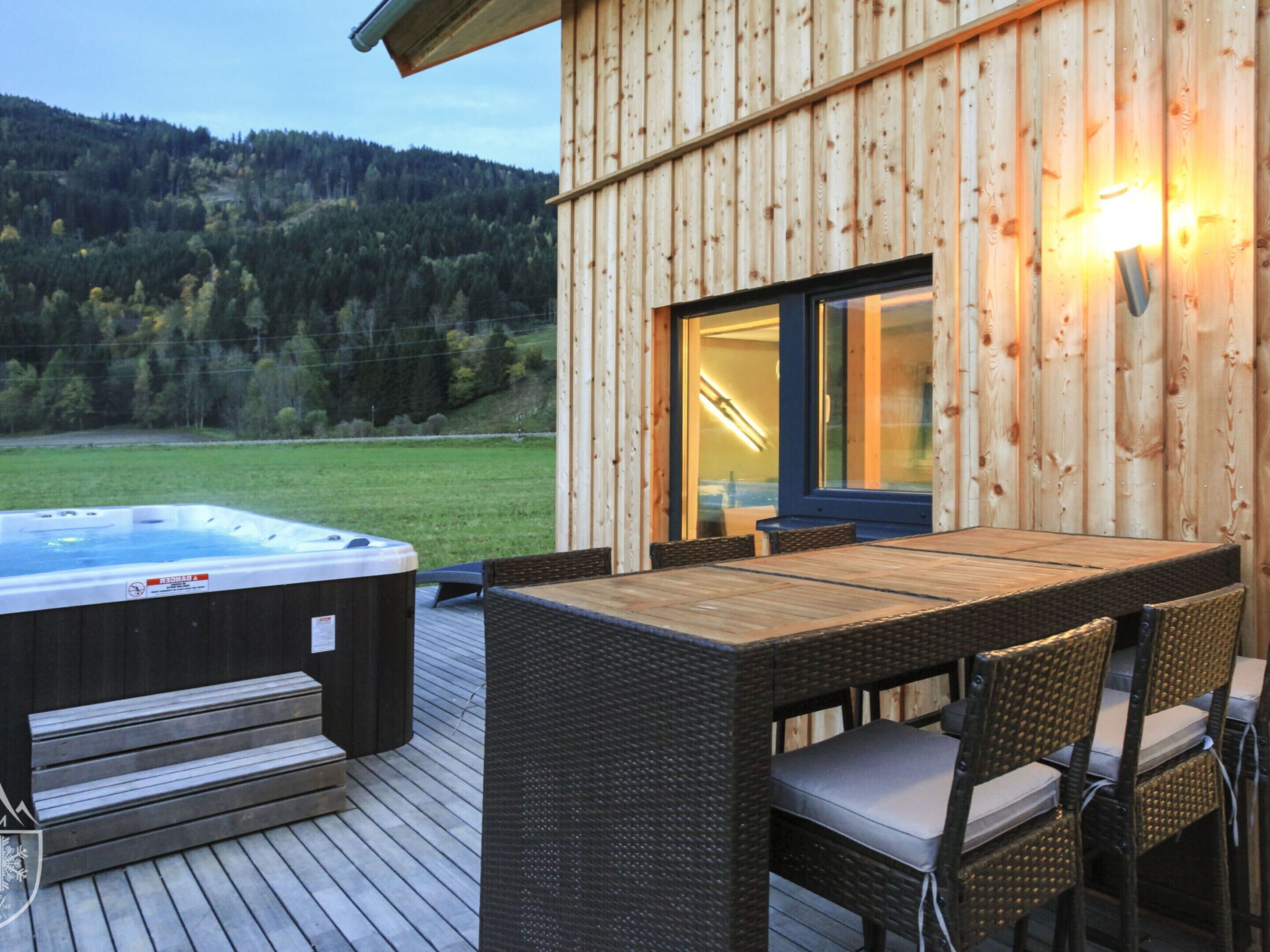 Una terraza de madera con un jacuzzi, mobiliario de mimbre y vistas a una ladera boscosa.