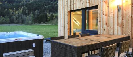 Una terraza de madera con un jacuzzi, mobiliario de mimbre y vistas a una ladera boscosa.