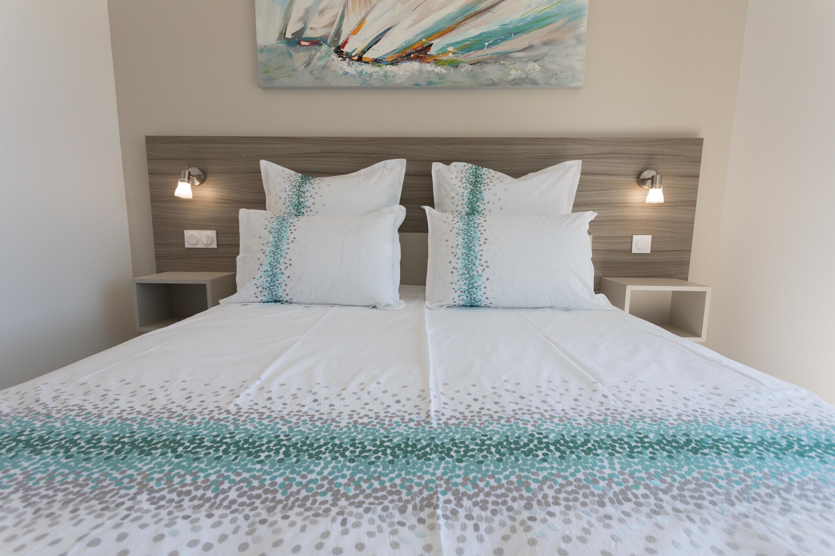 A double bed with a white and teal patterned bedspread, two white pillows with teal accents, wooden headboard, and two bedside tables with wall-mounted lamps.