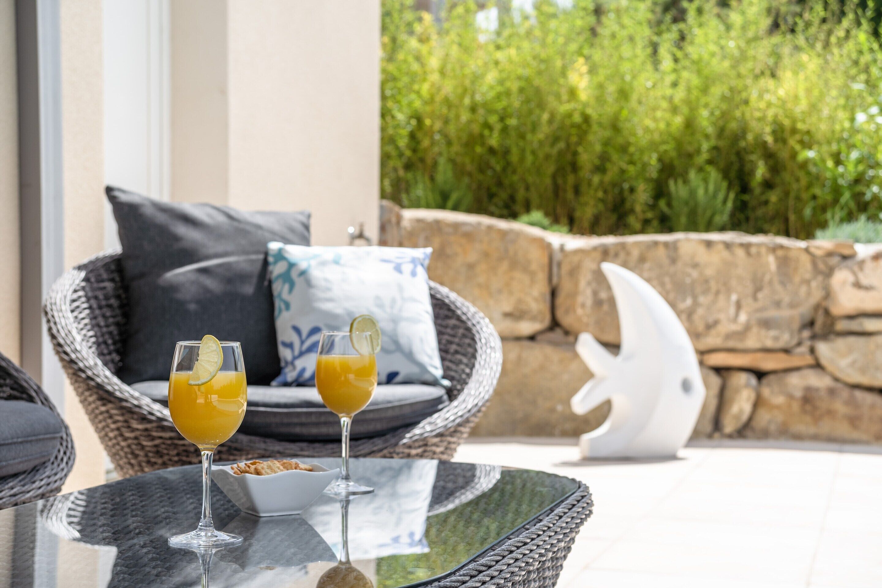 Two drinks on a glass table, wicker chairs with cushions, and a stone wall with greenery in the background.