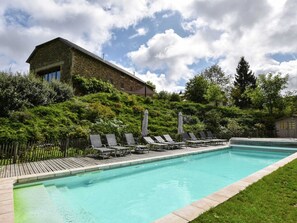 Una piscina amb gandules i ombrelles, envoltada de vegetació exuberant i un edifici de pedra.