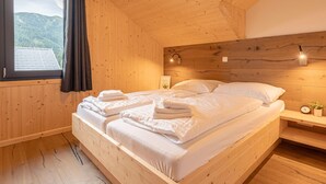 A wooden cabin bedroom with a bed, a nightstand, a wall-mounted lamp, and a window with curtains. - Premium Chalet With Terrace (Hohentauern)