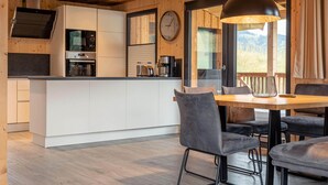 A modern kitchen with white cabinets, a built-in oven, and a coffee machine. There is a dining area with a wooden table and grey upholstered chairs. - Premium Chalet With Terrace (Hohentauern)