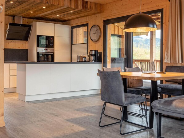 Une cuisine moderne avec des meubles blancs, un four encastré et une machine à café. On y trouve également un coin repas avec une table en bois et des chaises à revêtement gris.