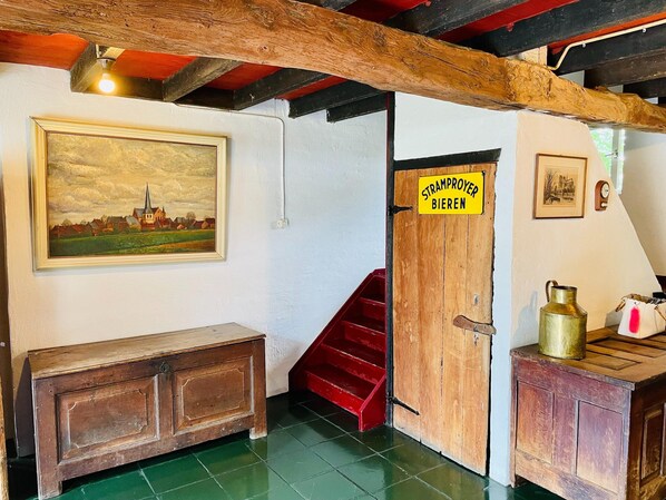 A room with a wooden bench, a staircase, a door labeled 'STRAMPROYER BIEREN,' a framed painting, and a brass pitcher on a wooden cabinet.