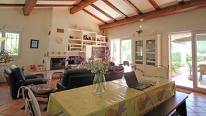 A living room with a dining table, chairs, a sofa, a fireplace, shelves with books, and a large window. - Villa in Bagnols Near Sea With Pool (Bagnols-en-Foret)