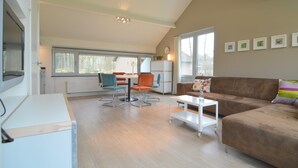 En rummelig opholdsstue med en brun lædersofa, et hvidt sofabord og orange lænestole. Der er et spisebord med stole, et hvidt køleskab og et vindue med persienner.