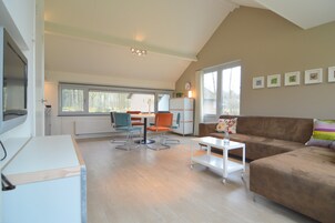 A spacious living area with a brown leather couch, a white coffee table, and orange chairs. There is a dining table with chairs, a white refrigerator, and a window with blinds.