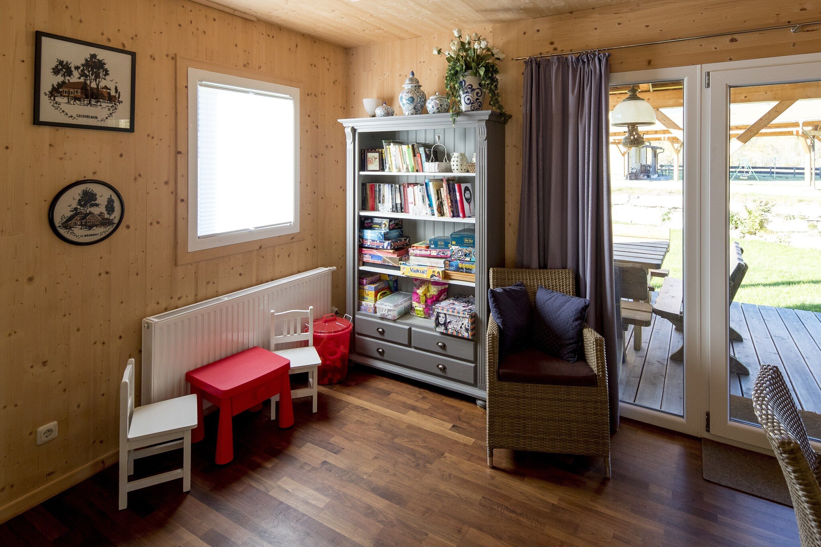 Ein gemütliches Zimmer mit Holzboden, einem roten Tisch, einem weißen Stuhl, einem Korbstuhl, einem Bücherregal und Blick durch eine Glastür.