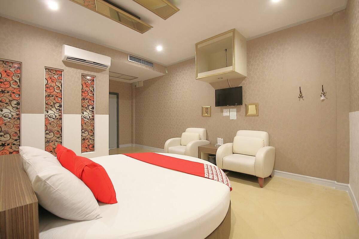 A hotel room with a large bed, two chairs, a TV, and an air conditioning unit.