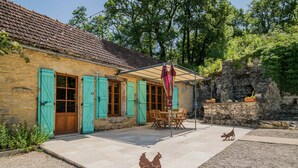 Ein Steinhaus mit Ziegeldach, einer Terrasse mit Tisch und Stühlen sowie einer Hühnerfigur.