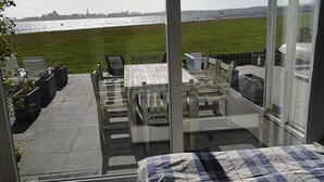 Ein Schlafzimmer mit großem Fenster, das auf eine Wiese und ein Gewässer blickt. Es gibt ein Bett mit karierter Decke, einen Holztisch mit Stühlen und eine Glastür, die auf eine Terrasse mit Grill führt.