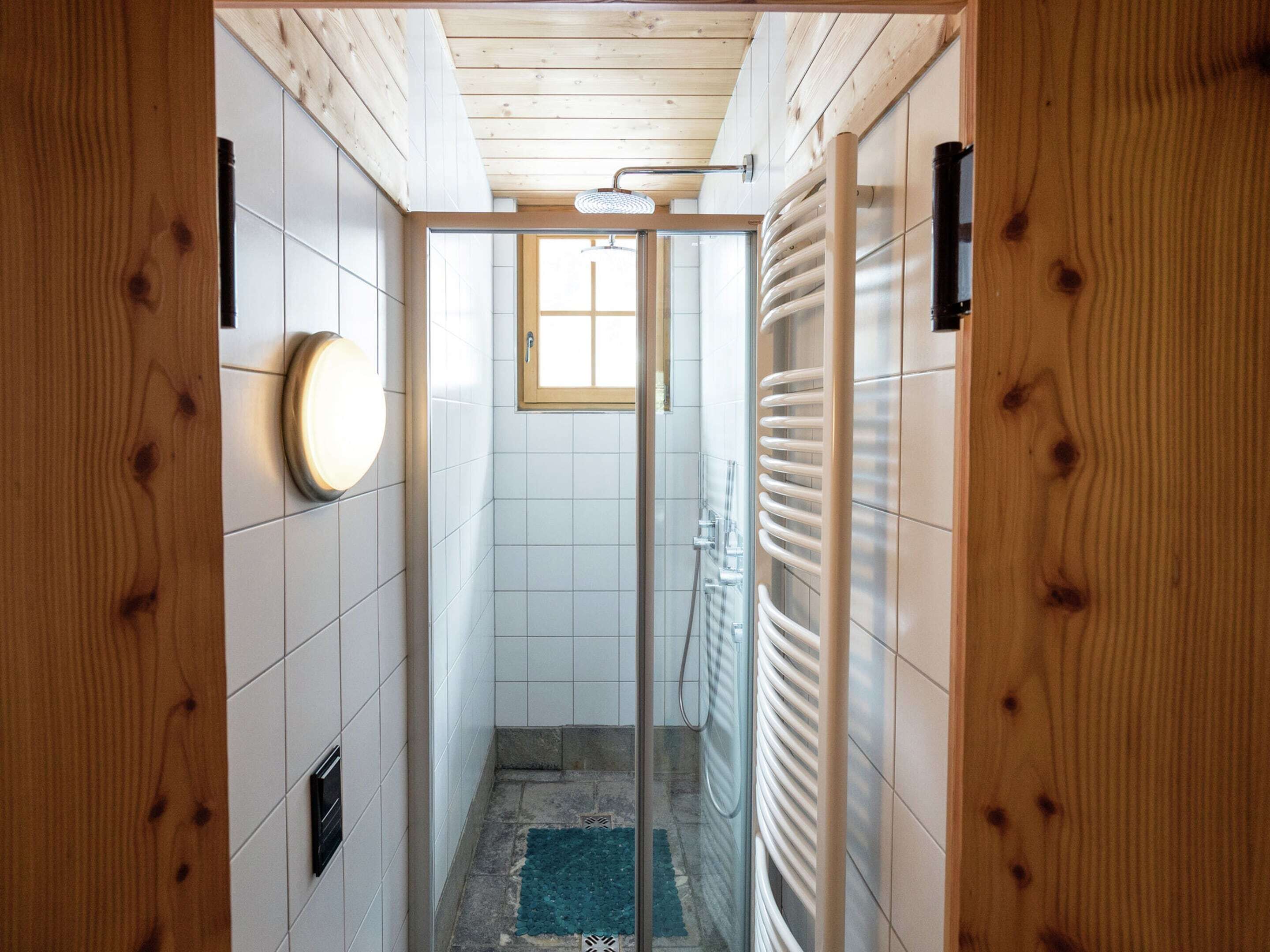 A narrow hallway with wooden walls, a tiled floor, a glass door leading to a shower area, and a small window.