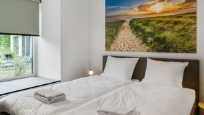 Une chambre avec un grand lit, une fenêtre et un tableau représentant un sentier de plage.