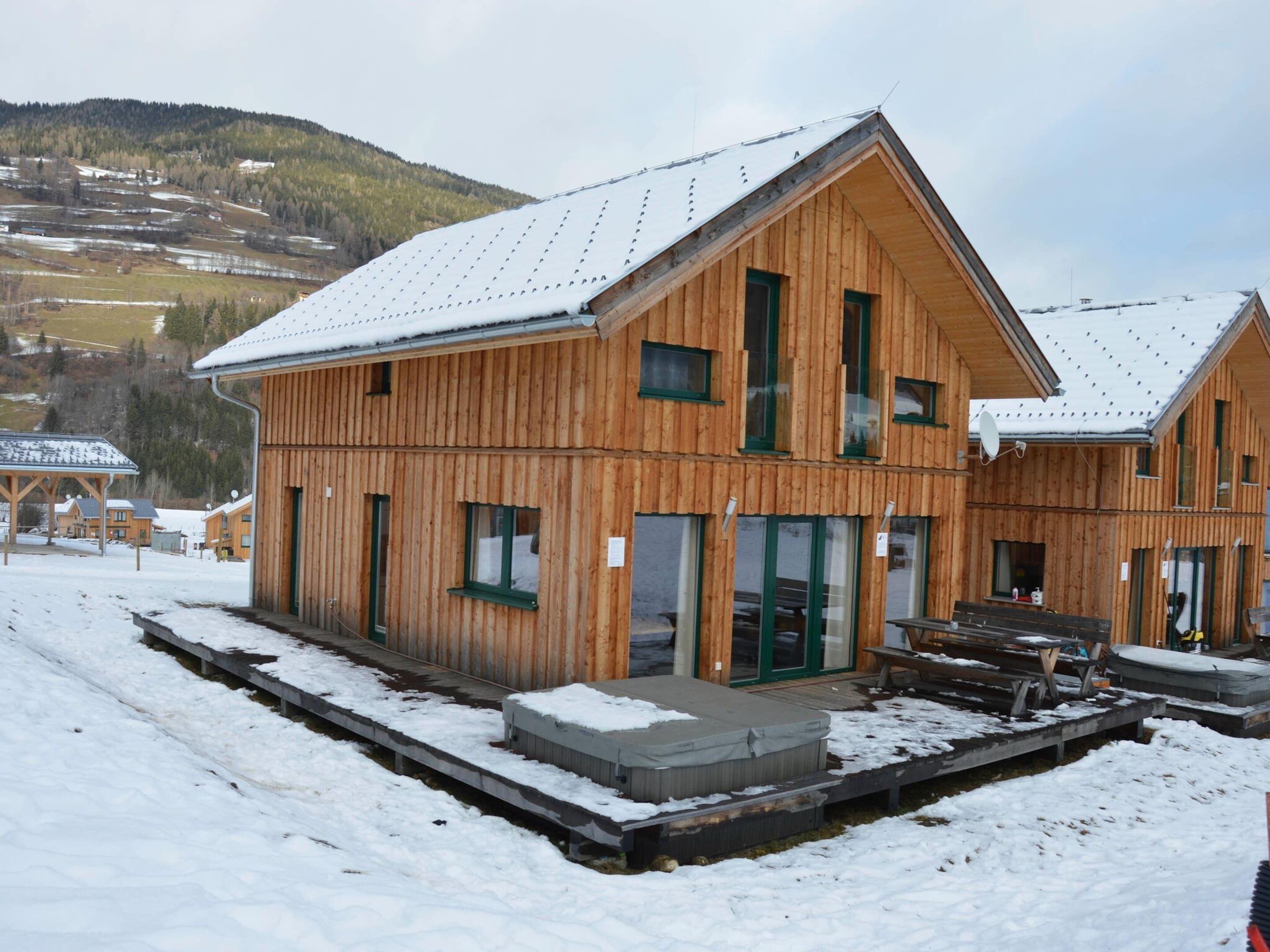 A wooden chalet with a snow-covered roof, a covered outdoor seating area, and a view of a mountainous landscape in the background.
