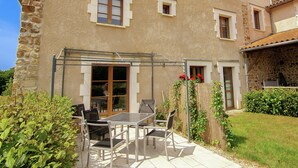 Un bâtiment à deux étages avec une façade en pierre, une entrée avec une porte en verre et un patio aménagé avec du mobilier d’extérieur.