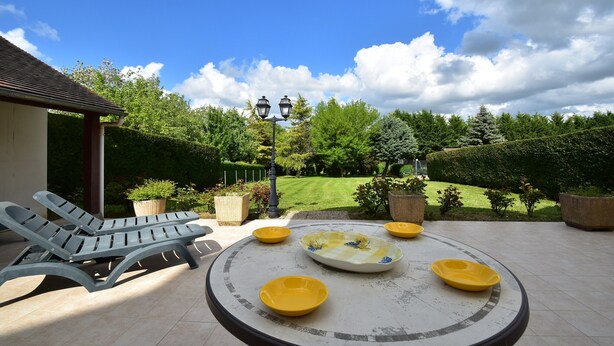 A patio with two lounge chairs, a round table with yellow bowls, and a view of a well-manicured garden and lawn. - Apartment in Faverolles Near the Beach (Villentrois-Faverolles-en-Berry)