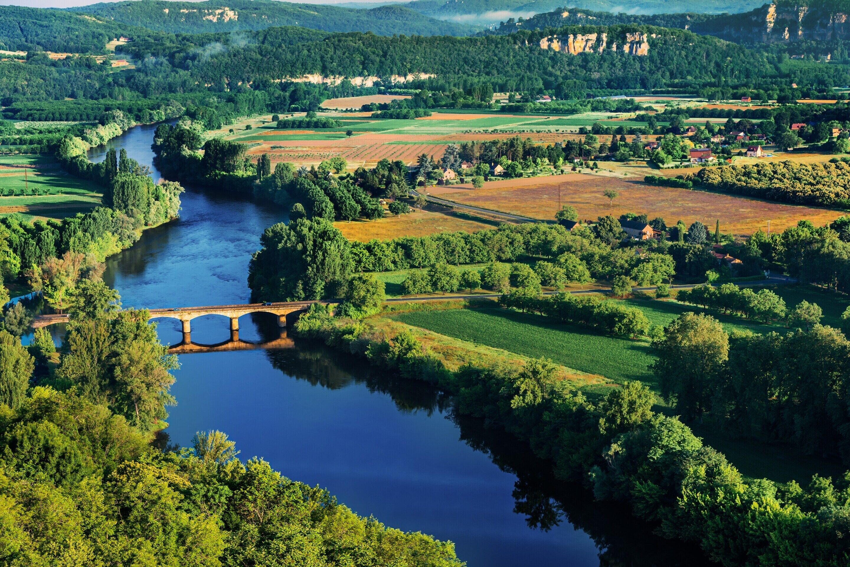 Um rio com uma ponte, rodeado por campos verdes e árvores.