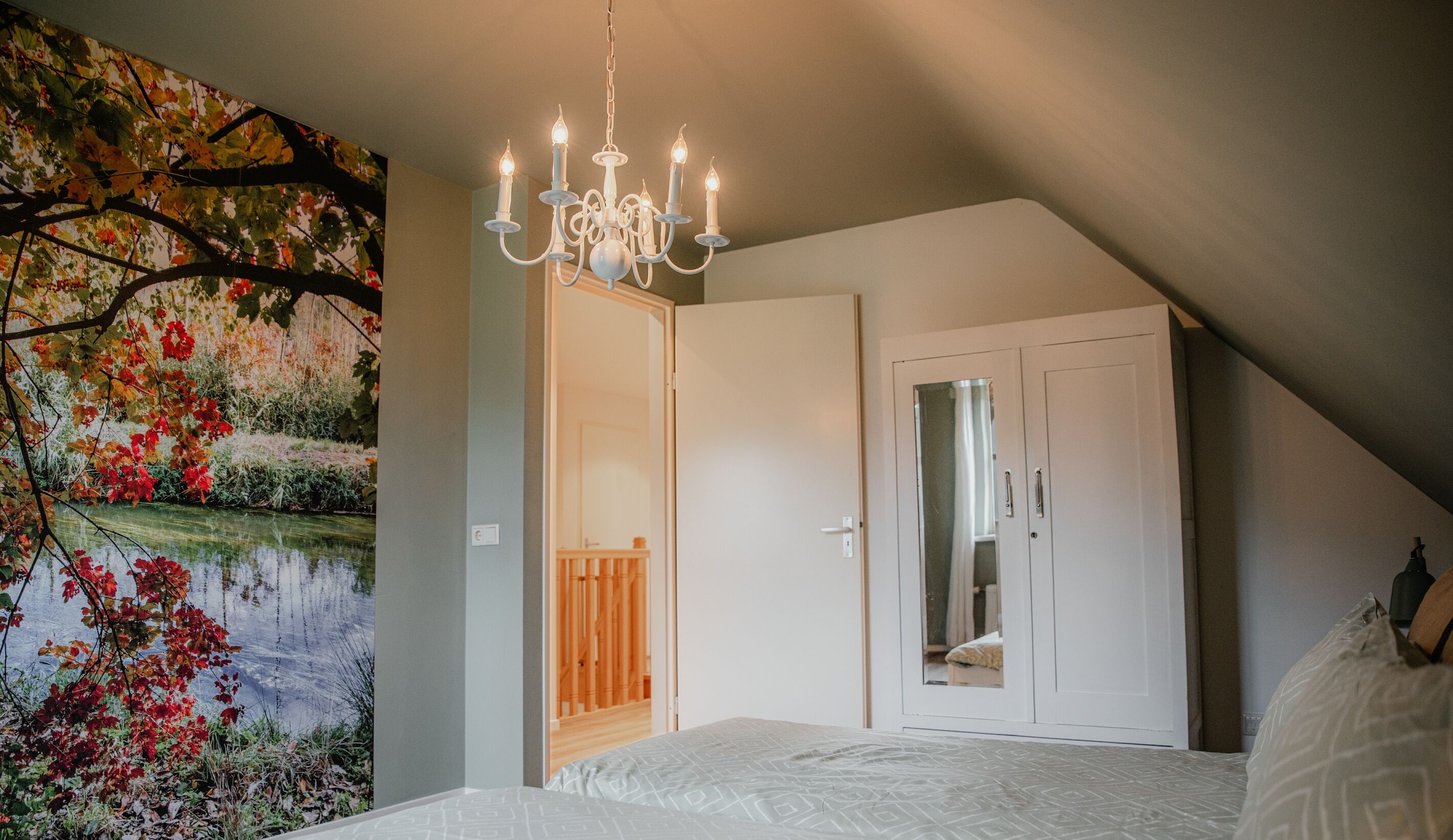A bedroom with a sloped ceiling, a chandelier, a bed with a patterned bedspread, a white wardrobe, and a wall with a nature-themed mural.