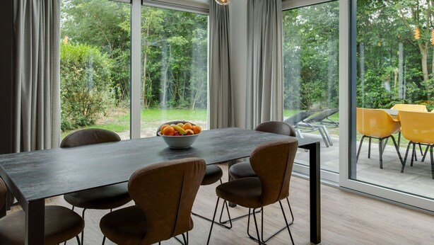 A modern dining area with a dark wooden table, brown upholstered chairs, and a bowl of fruit. Large windows offer a view of a lush garden and outdoor seating. - Bungalow in Texel Near National Park (De Koog)