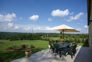 An outdoor patio with a table and chairs, a large umbrella, and a view of a green landscape. - Cosy Holiday Home Near the Causses du Quercy (Lavercantiere)