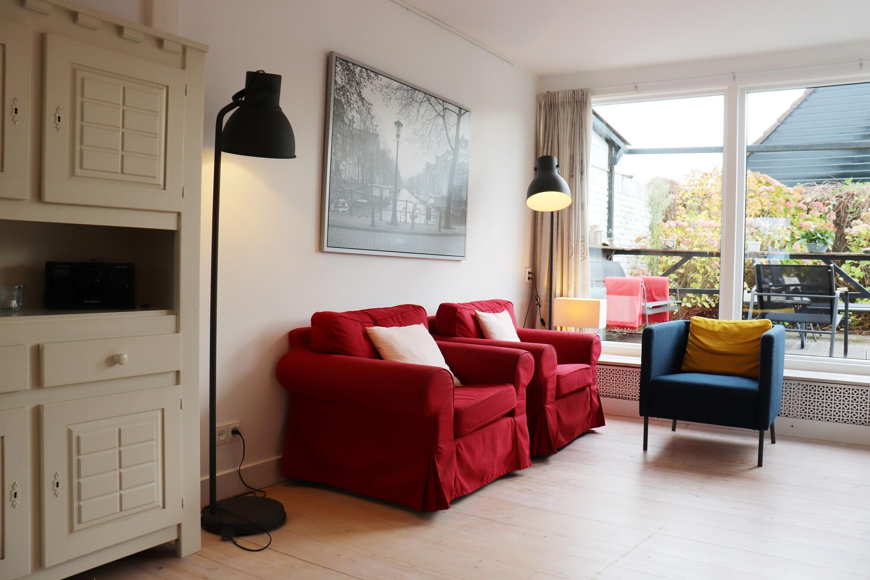 Una habitación con un sofá rojo, un sillón azul, un armario, una lámpara de pie y un ventanal con vistas al exterior.