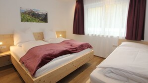 A bedroom with a wooden bed, white bedding, a nightstand, a wall-mounted lamp, a window with curtains, and a mountain landscape painting. - Luxurious Apartment in Fugenberg With Balcony (Fuegenberg)